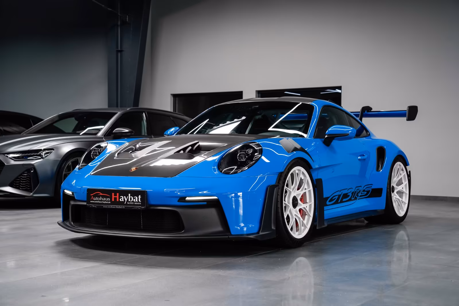 The Shark Blue GT3 RS at front three-quarter in a showroom, carbon bonnet vents and white wheels
